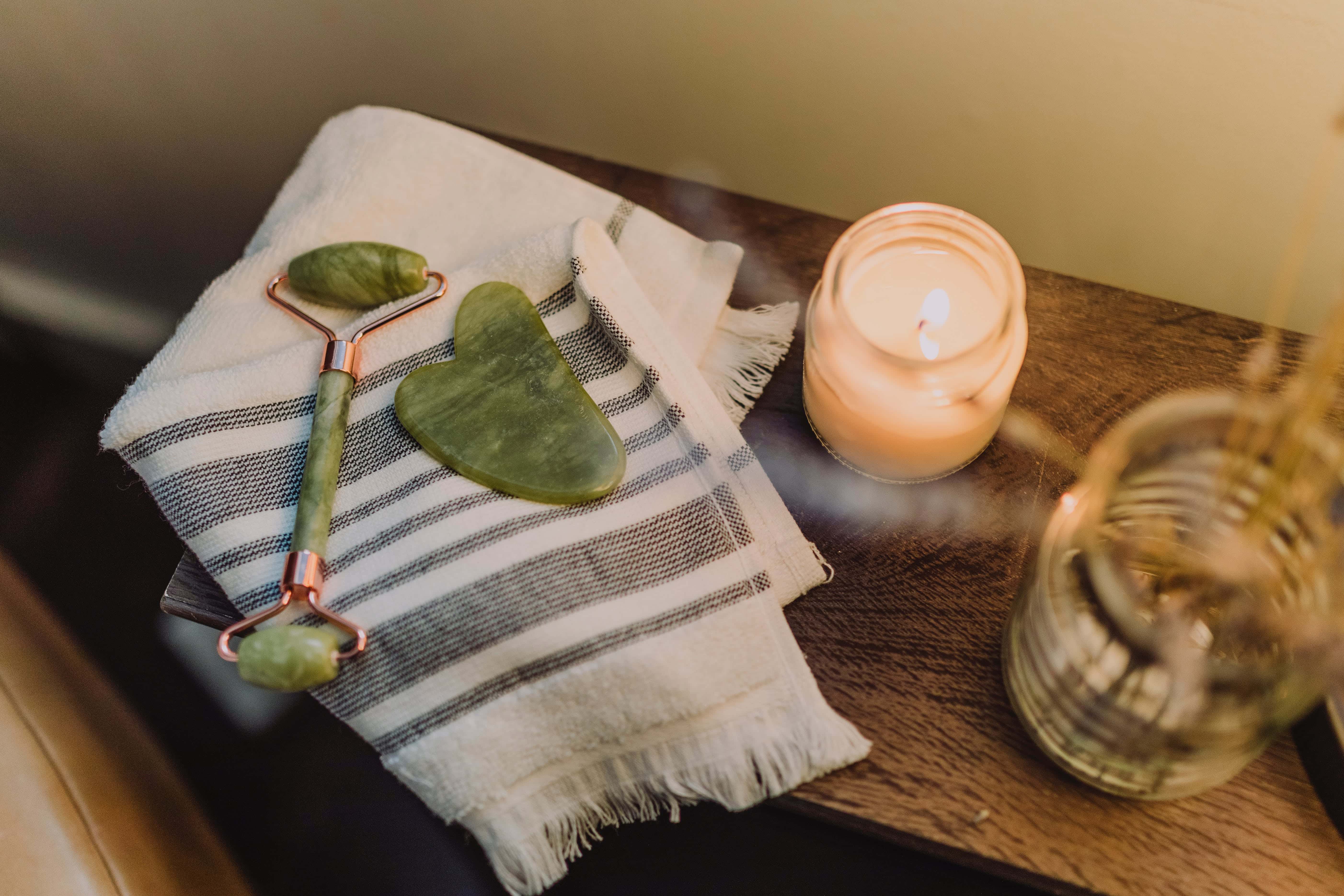 Close-up of a spa setting featuring a green jade facial roller and a heart-shaped gua sha tool on a striped towel, with a lit candle providing a warm glow