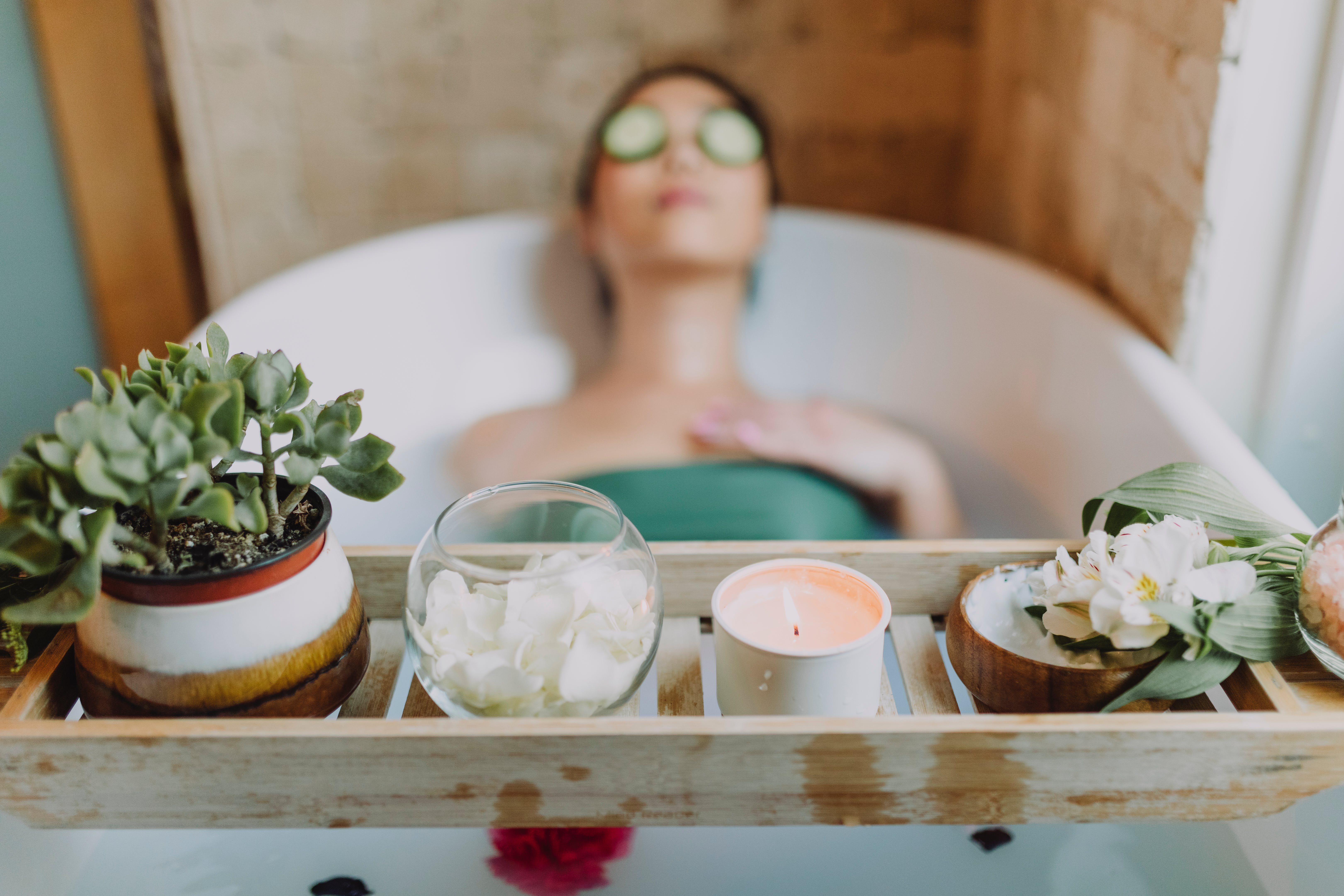 A pampered spa experience depicted by a woman reclining in a bath with cucumber slices over her eyes, surrounded by a wooden tray holding a candle, succulents, and a bowl of flower petals, indicative of the luxurious treatments available at the spa